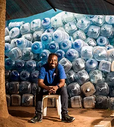 Man sitting on a stool in front of stacked water jugs. Click to learn more about Water & Sanitation.