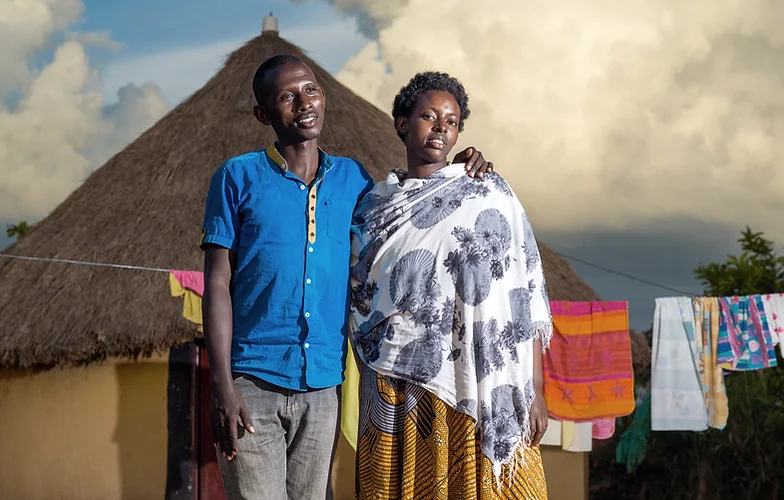 Couple standing together in front of a thatched house with laundry.